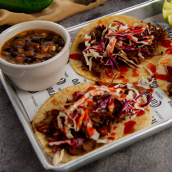 Two El Patron Mexican Grill tacos with shredded meat, cabbage, and sauce, served with a side of black beans.
